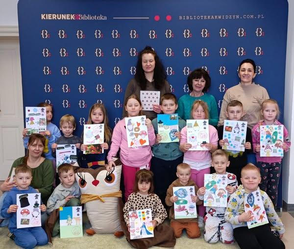 Zdjęcie przedstawia przedszkolaki, wychowawcę i opiekuna wycieczki wraz z pracownikami biblioteki. Na zdjęciu dzieci trzymają wykonane podczas warsztatów prace. 