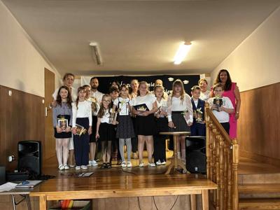 Zdjęcie grupowe uczniów kl. III wraz z dyrektorem szkoły, wychowawcą i rodzicami. Dzieci ubrane w galowym stroju stoją na scenie. W tle dekoracja z okazji zakończenia roku szkolnego. 