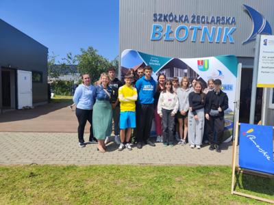 Na zdjęciu grupa uczniów z opiekunami/. Uczniowie stoją na tle budynku Szkółki Żeglarskiej. 