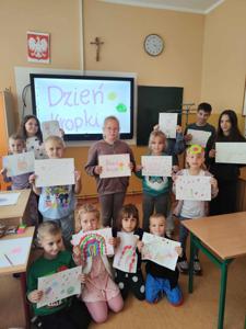 Na zdjęciu grupa dzieci podczas zajęć świetlicowych, trzymająca swoje prace plastyczne z wykorzystaniem kropek. Tło - tablica multimedialna.