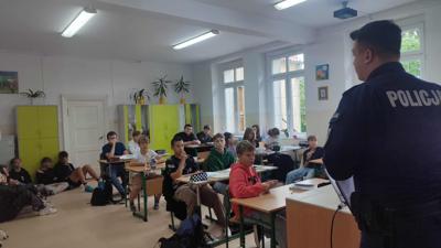 Na zdjęciu funkcjonariusz policji prowadzący zajęcia dla uczniów. Dzieci siedzące w ławkach, podnoszą ręce, zgłaszają się . W tle widać szkolne wyposażenie sali lekcyjnej. 