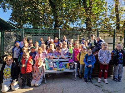 Na zdjęciu widzimy dużą grupę dzieci, które stoją razem na tle ogrodzenia i drzew w słoneczny dzień. Dzieci są uśmiechnięte, ubrane kolorowo – część ma kurtki, bluzy, a niektóre plecaki. Przed nimi stoi biała ławka, na której ustawione są  paczki karmy, zabawki i akcesoria dla zwierząt. Wygląda na to, że dzieci przyszły z wizytą do schroniska dla zwierząt, przynosząc ze sobą dary dla podopiecznych. Atmosfera na zdjęciu jest radosna i przyjazna.