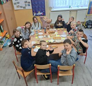 Uczniowie klasy trzeciej siedzący wokół szkolnych ławek. Na ławkach ułożone są różne rodzaje pieczywa oraz mąk, kasz i płatków.