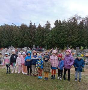 Na zdjęciu widać grupę dzieci stojących w rzędzie na tle cmentarza. Za nimi widoczne są groby ozdobione kwiatami i zniczami oraz rząd wysokich, zielonych drzew iglastych. Każde dziecko ubrane jest kolorowo, w różne wzory i odcienie. Scena wygląda spokojnie i zorganizowanie, jakby dzieci przyszły tu w ramach szkolnej lub przedszkolnej wizyty, być może z okazji dnia pamięci lub odwiedzin grobów.