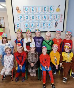 Na zdjęciu widać grupę dzieci przebranych w kolorowe stroje z bajek i filmów animowanych. Siedzą i stoją w dwóch rzędach przed tablicą z napisem „DZIEŃ POSTACI Z BAJEK”. Dzieci mają na głowach papierowe opaski ozdobione kolorowymi rysunkami. Ich kostiumy przedstawiają różne bajkowe postacie. W tle widać ilustracje bohaterów z bajek, takich jak Kubuś Puchatek, Królewna Śnieżka i inni. Zdjęcie ma radosny, kolorowy i przyjazny klimat — widać, że to przedszkolne świętowanie.