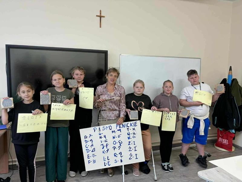 Na zdjęciu uczniowie klasy 5 wraz z nauczycielem historii, Panią Anną Kowalczyk. Uczniowie trzymają w dłoniach gliniane tabliczki z pismem klinowym oraz wzór pisma supełkowego i wzór hieroglifów.