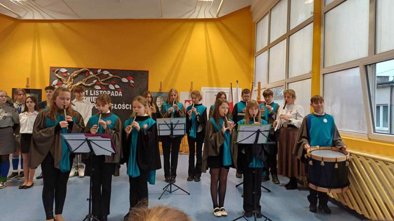 Występ zespołu Flauto Dolce, który gra na flażoletach. Po prawej stronie uczeń gra na tarabanie, obok niego gra nauczyciel. Wszyscy ubrani są w togi. 