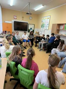 Na zdjęciu widać grupę uczniów siedzących w kręgu w szkolnej sali podczas warsztatów profilaktycznych. Dzieci słuchają prowadzącej, która znajduje się w środku kręgu. W tle widoczna jest tablica multimedialna, zegar oraz kolorowe prace plastyczne na ścianach. Atmosfera jest spokojna i sprzyjająca rozmowie.
