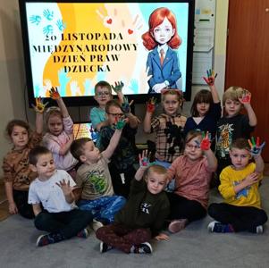 Na zdjęciu widać grupę dzieci siedzących na dywanie w sali przedszkolnej. Dzieci trzymają w górze dłonie pomalowane farbami w różnych kolorach. Wszystkie są uśmiechnięte i skupione. Za nimi stoi duży ekran multimedialny. Na ekranie widnieje kolorowa grafika z napisem: „20 LISTOPADA MIĘDZYNARODOWY DZIEŃ PRAW DZIECKA”. Cała scena przedstawia obchody Międzynarodowego Dnia Praw Dziecka — dzieci biorą udział w aktywności artystycznej podkreślającej znaczenie praw dziecka.