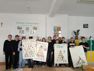 Na zdjęciu widać grupę uczniów i nauczycielkę stojących w klasie. Trzymają plakaty przedstawiające piramidę zdrowego żywienia. Wszyscy wyglądają na zaangażowanych w projekt edukacyjny.