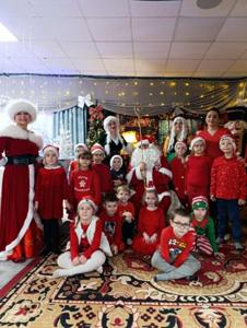 Na zdjęciu widać dużą, świąteczną grupę dzieci i dorosłych pozujących do wspólnej fotografii. Scena jest utrzymana w bożonarodzeniowym klimacie: dzieci mają na sobie czerwone stroje, sukienki i świąteczne czapki, a część z nich siedzi na dywanie, inni stoją w rzędach. W centrum znajduje się osoba przebrana za Świętego Mikołaja z laską.